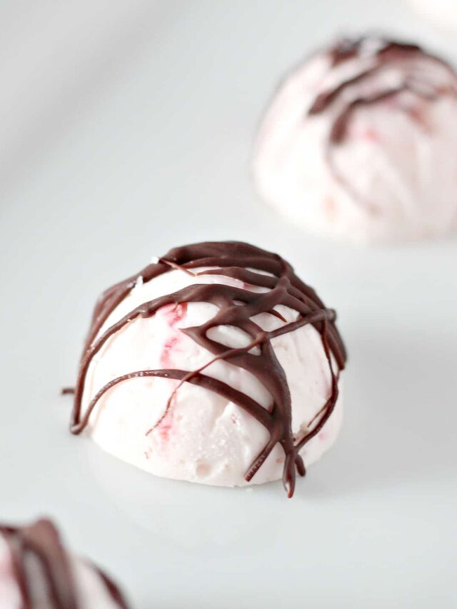 Close-up of a round, white fat bomb dessert with pink strawberry swirls, topped with thin drizzles of chocolate. The dessert appears to have a creamy texture and is placed on a white surface.