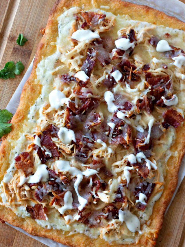 A crispy rectangular keto pizza topped with shredded chicken, bacon bits, and drizzles of white sauce on a wooden surface, garnished with cilantro on the side.