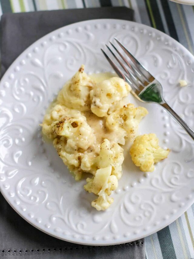 A white, ornate plate holds a serving of cheesy cauliflower bake with a creamy sauce, accompanied by a fork resting on the edge.