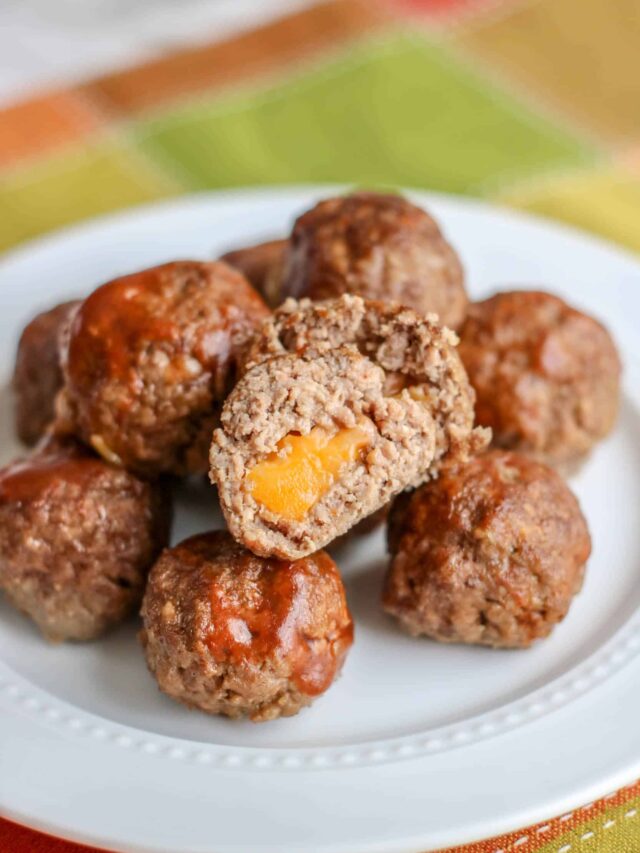 Plate of easy, cheese-stuffed barbecue meatballs with one cut open to reveal the gooey center. White plate set on a multicolored tablecloth.