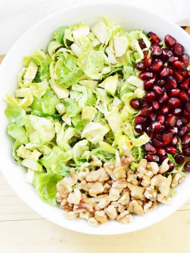 A bowl of salad with chopped brussels sprouts, pomegranate seeds, and walnuts, separated into sections on a white background.