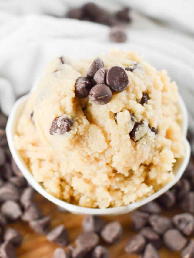 A bowl of low-carb chocolate chip cookie dough, topped with several chocolate chips, with more chocolate chips scattered around on a wooden surface.