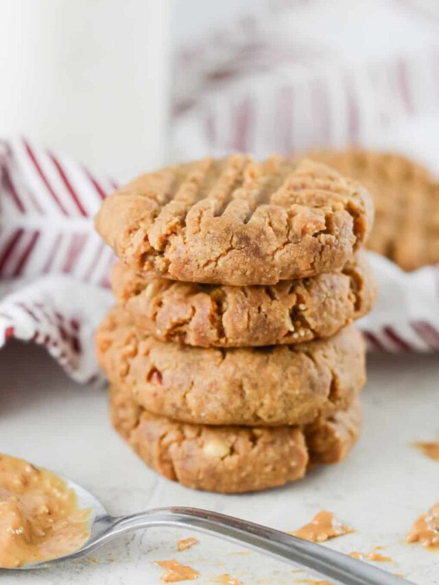 A stack of four keto peanut butter cookies is placed in front of a striped cloth, with a spoon of rich peanut butter in the foreground.