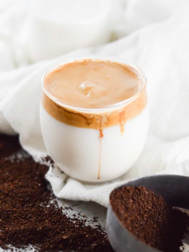 A glass of Dalgona coffee with creamy coffee foam on top, placed on a white cloth background with coffee grounds in a bowl nearby, perfectly illustrating the dalgona coffee recipe.