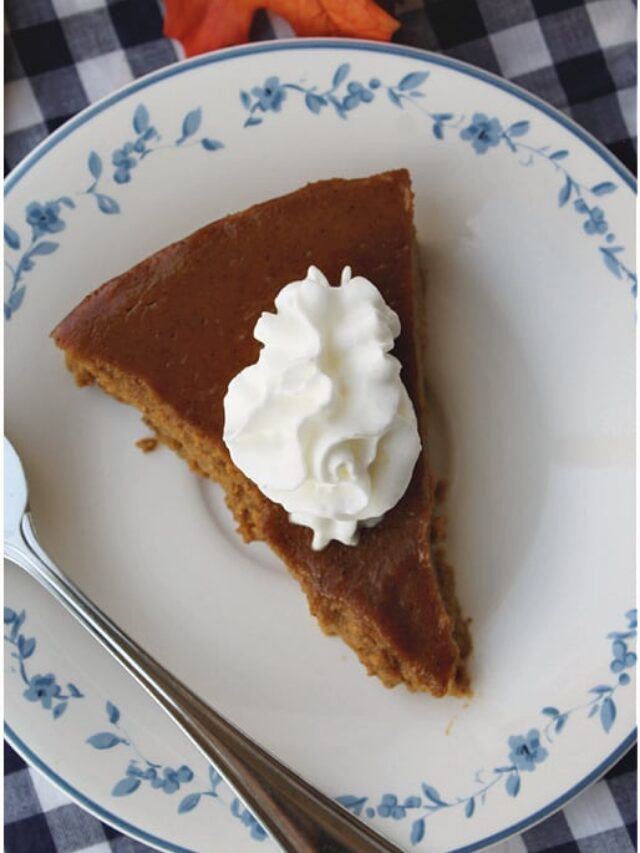 A slice of keto-friendly pumpkin pie topped with whipped cream on a floral plate, accompanied by a spoon, set on a checkered tablecloth with a fall leaf in the background.