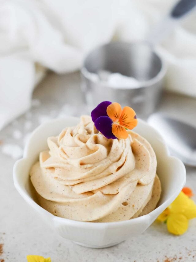 A bowl of swirled whipped cream mousse topped with two edible flowers—one purple and one orange—sits on a light-colored surface, with a spoon and ingredient container in the background.