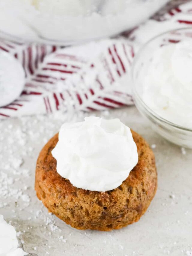 A small keto pastry topped with a dollop of white whipped cream rests on a white surface. In the background, a bowl of whipped cream and a red-patterned cloth complete the scene.