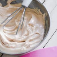A silver mixing bowl filled with thick, creamy homemade keto frosting being mixed by an electric hand mixer with white beaters.