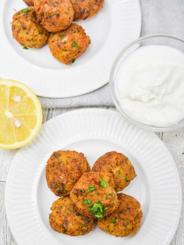 Two plates of cooked falafel balls garnished with herbs, accompanied by a lemon half and a bowl of white sauce, placed on a light-colored surface. These keto-friendly bites make for a delicious appetizer.