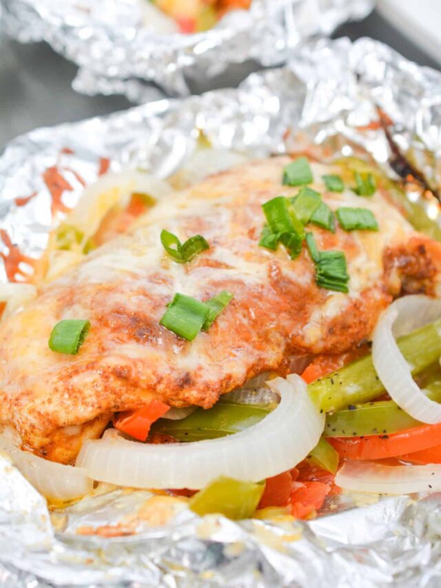 A foil packet containing a cooked piece of chicken breast topped with melted cheese and garnished with chopped green onions, resting on a bed of sliced onions and bell peppers, perfect for a Keto-friendly Tex Mex meal.