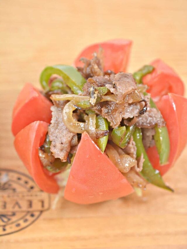 A dish featuring sliced beef, bell peppers, and onions is served within a carved tomato on a wooden surface. Perfect for those following a keto lifestyle or craving a Philly cheesesteak twist.