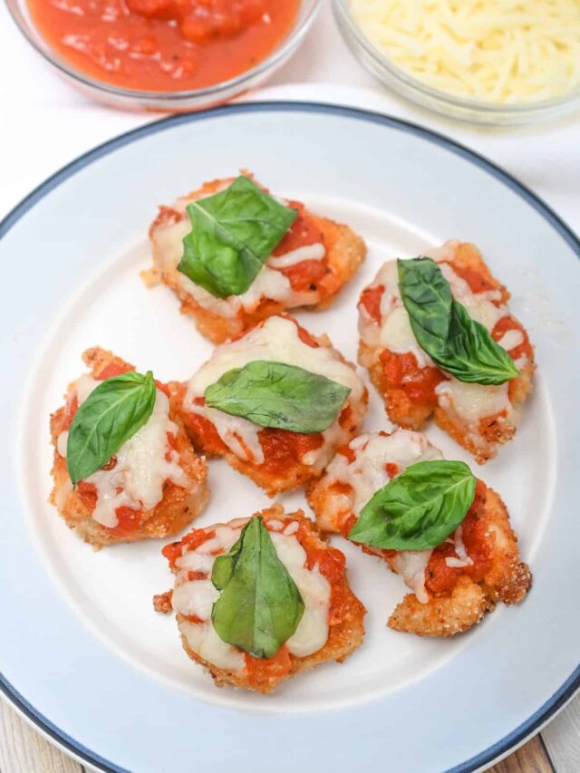 Plate with six breaded, bite-sized chicken appetizers topped with melted cheese, tomato sauce, and fresh basil leaves. Additional bowls containing tomato sauce and shredded parmesan cheese are in the background.