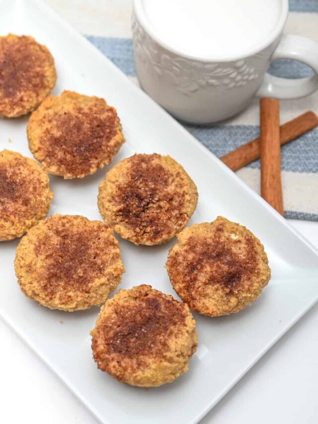 A rectangular white plate with eight keto cinnamon sugar mini muffins sits beside a white mug filled with milk and two cinnamon sticks on a blue and white striped cloth.