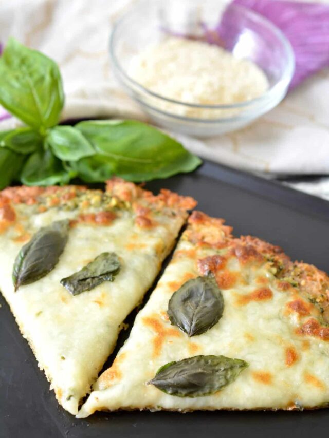 Two slices of low-carb pizza with melted cheese and basil leaves on a black plate, garnished with fresh basil. A small bowl of grated cheese is in the background.