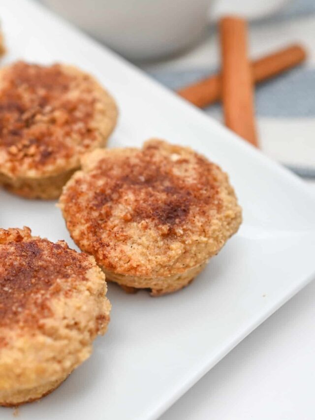 A close-up of mini Keto muffins on a white plate. The muffins have a crumbly, cinnamon-speckled topping. A pair of cinnamon sticks is visible in the background.