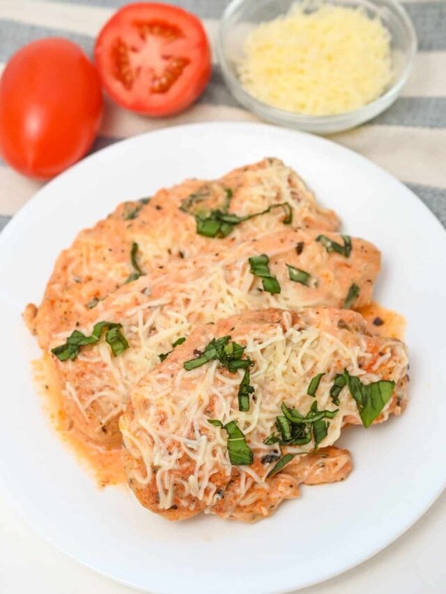 A plate of chicken parmesan topped with melted cheese and fresh basil, with a sliced tomato and grated cheese in the background.