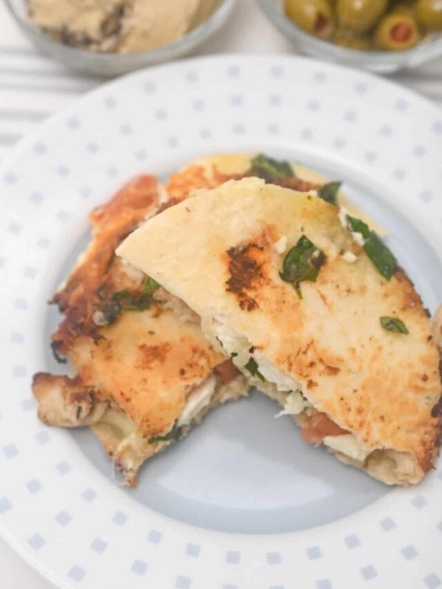 Two folded quesadillas with cheese and spinach on a white plate, accompanied by a blurred background of Mediterranean olives and another dish.