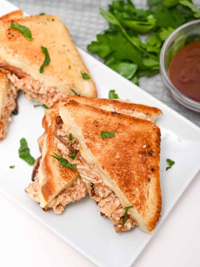 Toasted sandwich cut diagonally on a white plate, filled with tuna and garnished with parsley, served next to a small bowl of sauce.