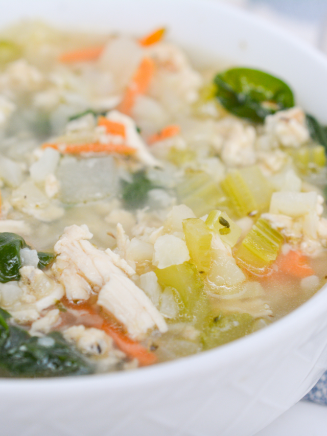 A close-up photo of a bowl of keto chicken and rice soup. The soup is yellow and has pieces of chicken, cauliflower rice, and carrots. There is a green garnish on top of the soup.