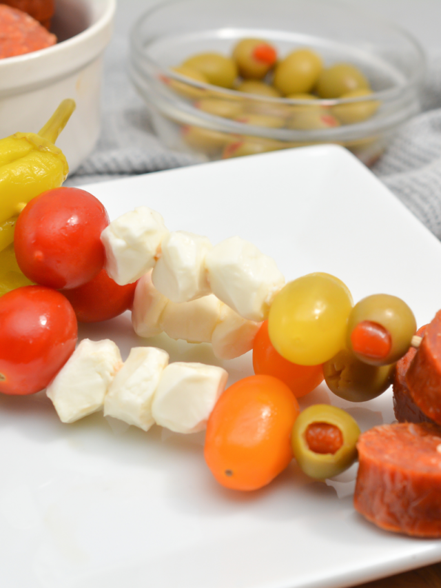 A plate with antipasto skewers holding tomatoes, cheese cubes, olives, and slices of sausage. A bowl with more olives is in the background.