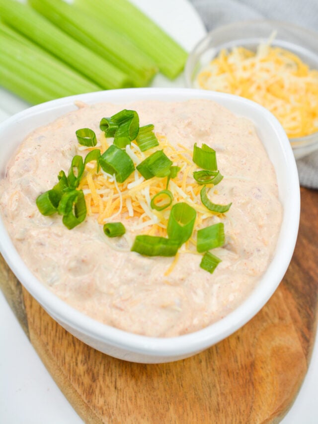 A bowl of creamy, keto-friendly dip topped with shredded cheese and chopped green onions sits on a wooden board with celery sticks. In the background, a small dish of shredded cheese completes this viral TikTok favorite known as boat dip.